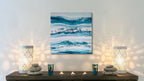 Abstract painting on a wall with two lit candles and decorative stones on a shelf below.