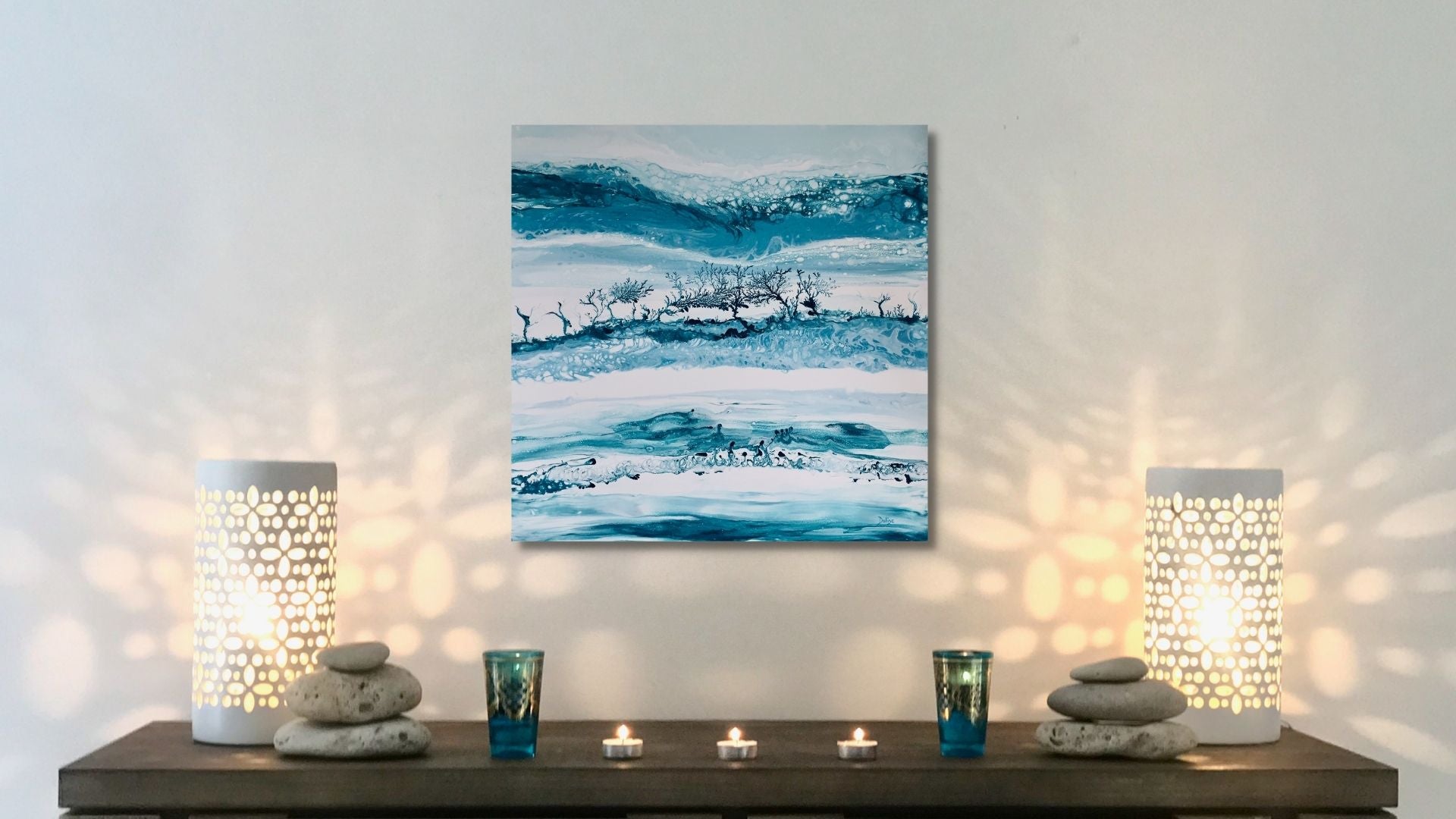 Abstract painting on a wall with two lit candles and decorative stones on a shelf below.